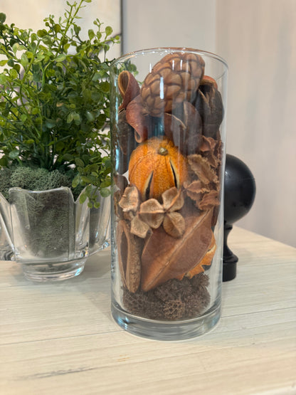 Glass vase with dried flowers and an orange on a wooden surface with a plant in the background