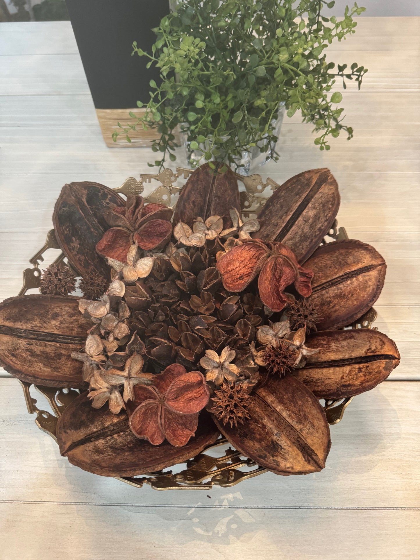 Decorative arrangement of dried flowers and leaves in a metal holder on a light wooden surface.