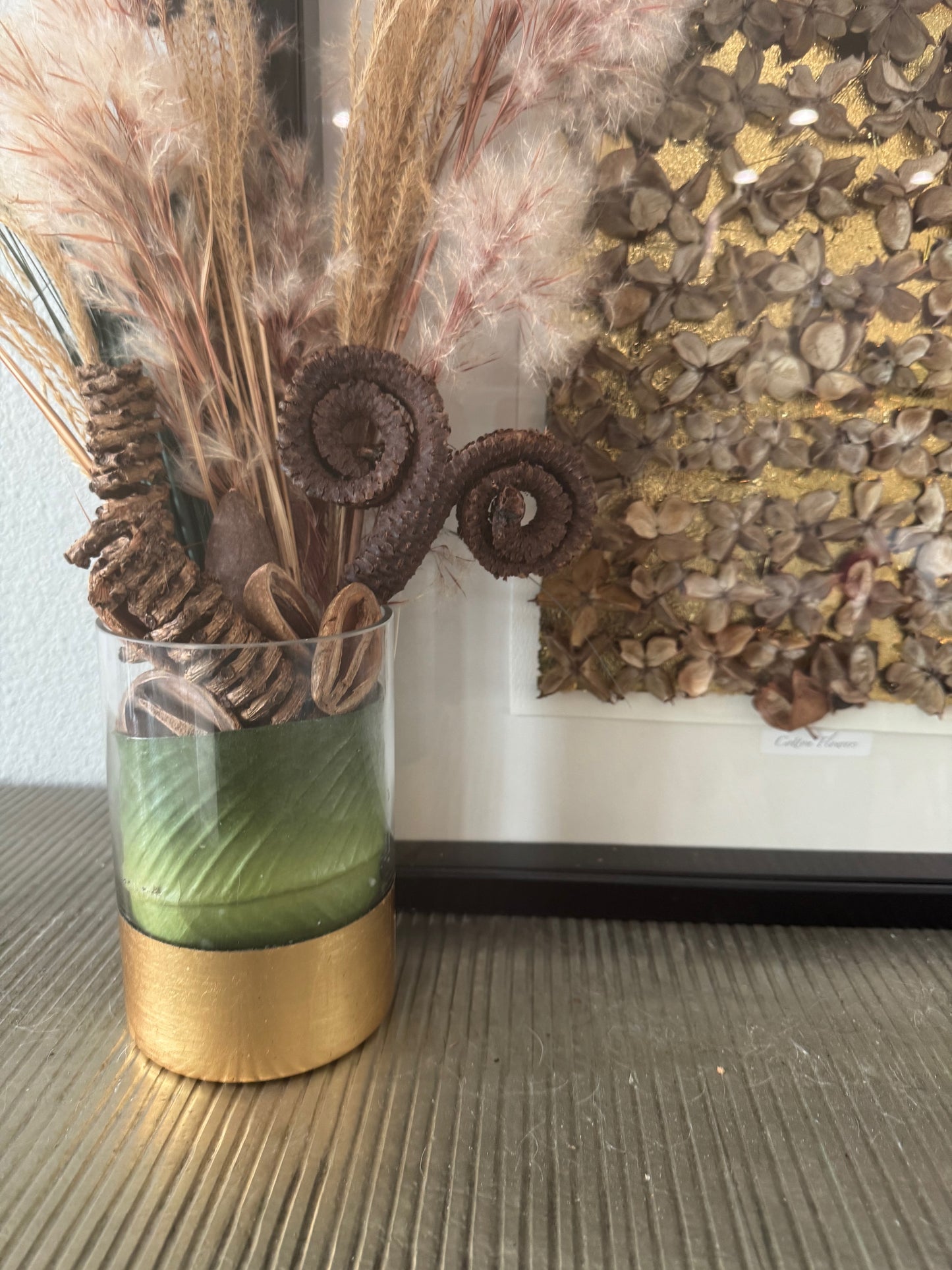 Decorative glass vase with gold base, green leaf, seed pods, and dried plants against a textured wall.