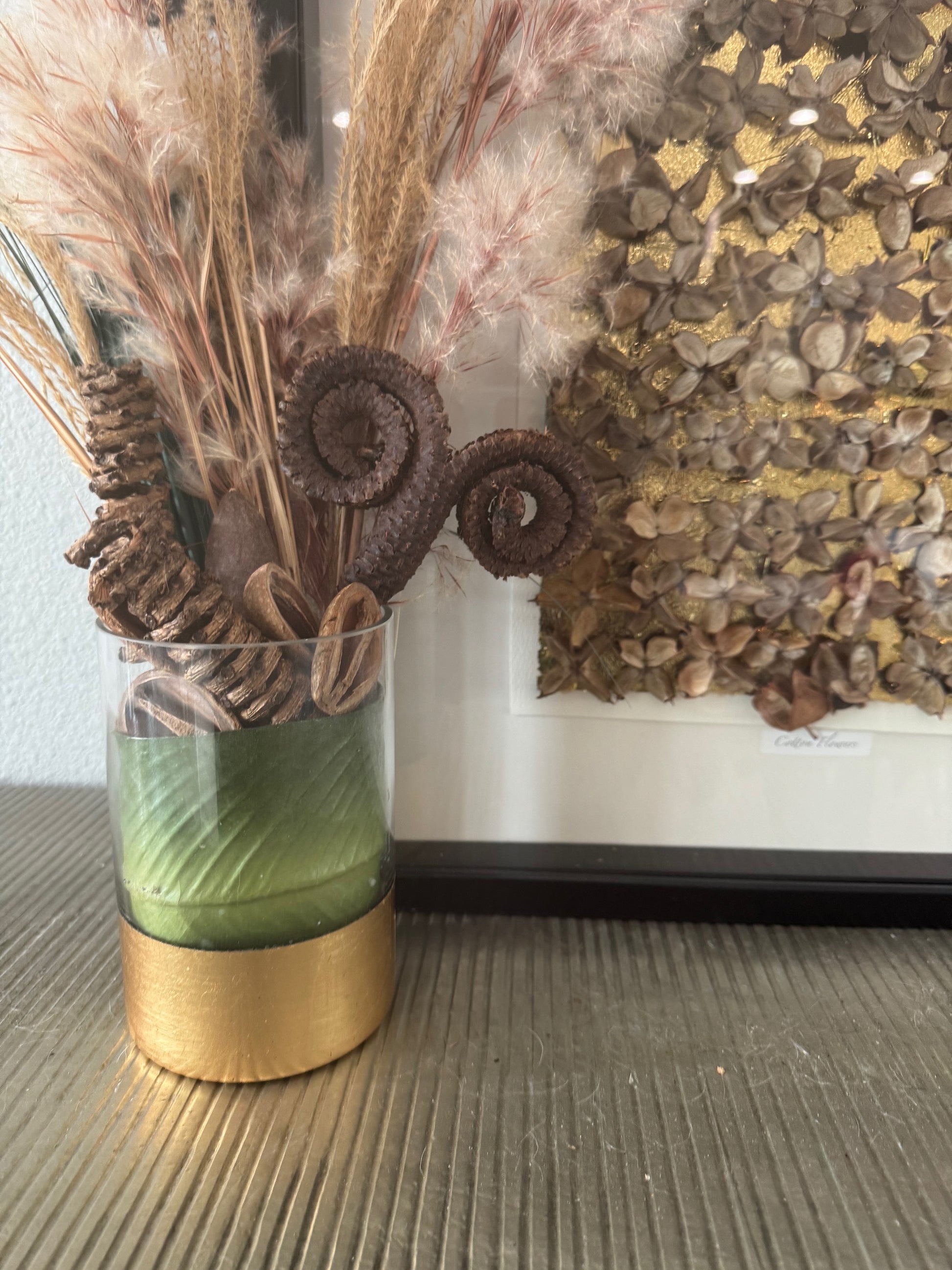 Decorative glass vase with gold base, green leaf, seed pods, and dried plants against a textured wall.