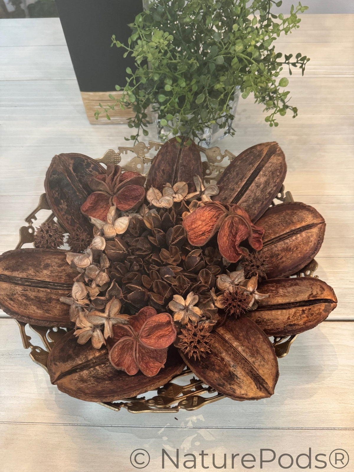 Decorative arrangement of dried flowers and leaves in a metal holder on a light wooden surface.